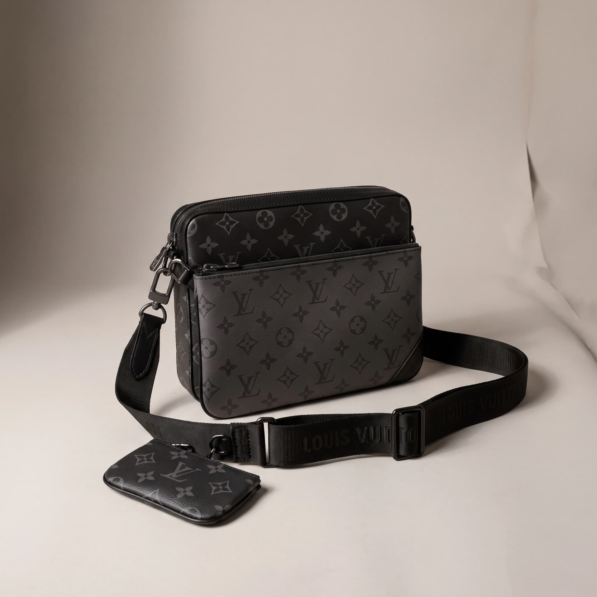 Trio Messenger en toile Monogram Eclipse de trois quarts, la pochette accrochée à la bandoulière posée devant.