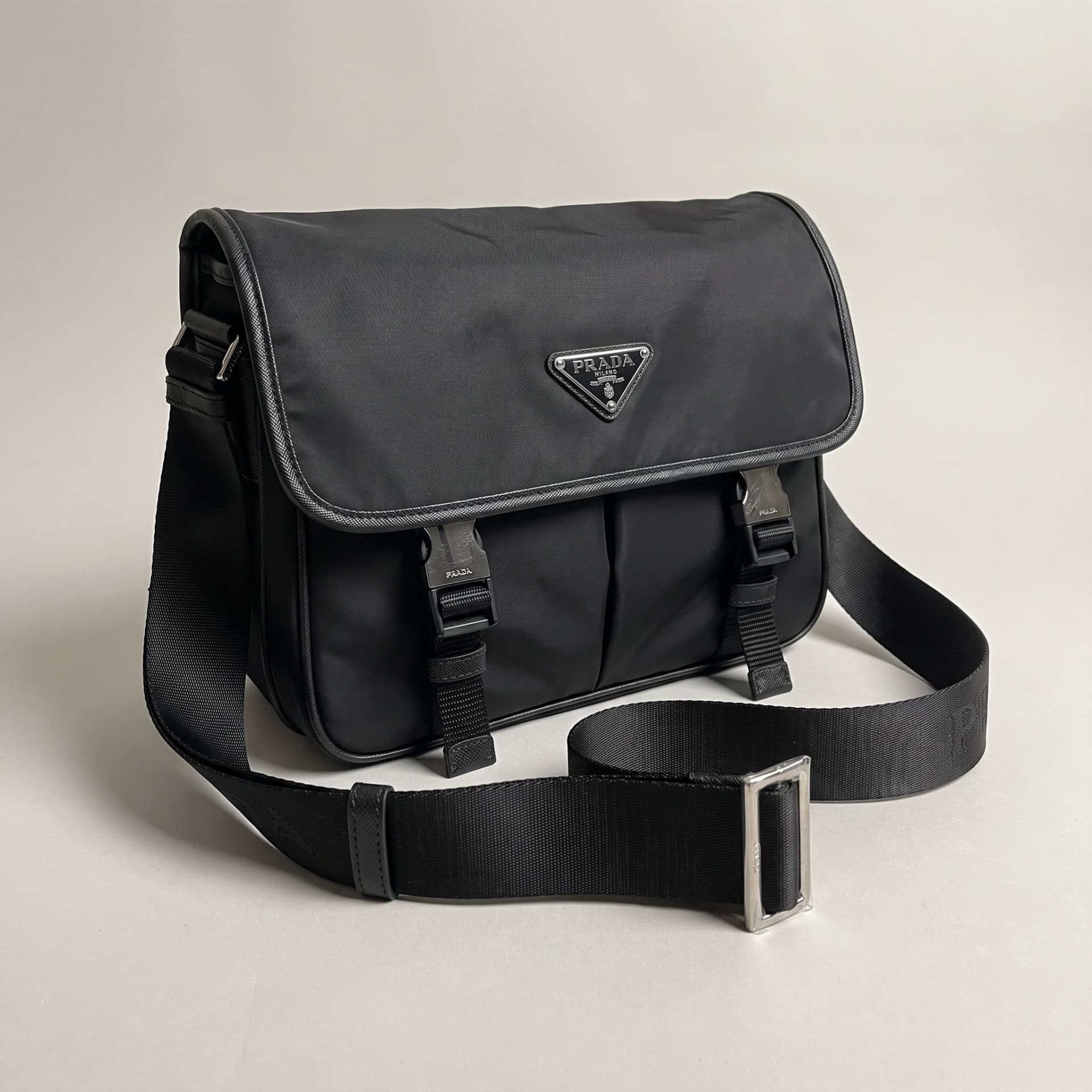 Messenger Prada Re-Nylon noir debout de trois quarts, rabat fermé par deux boucles, bandoulière déployée.